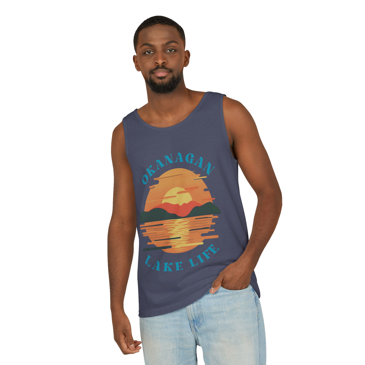 A man wearing a slate blue tank top with a colorful graphic of a sunset over water and the text 'OKANAGAN LAKE LIFE', paired with light wash jeans, set against a plain white background.