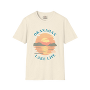 An off-white short-sleeve t-shirt with a retro-style graphic of a sunset over a lake, featuring the text 'OKANAGAN LAKE LIFE' in blue. The brand 'XXX Design' is visible on the inner neck label.