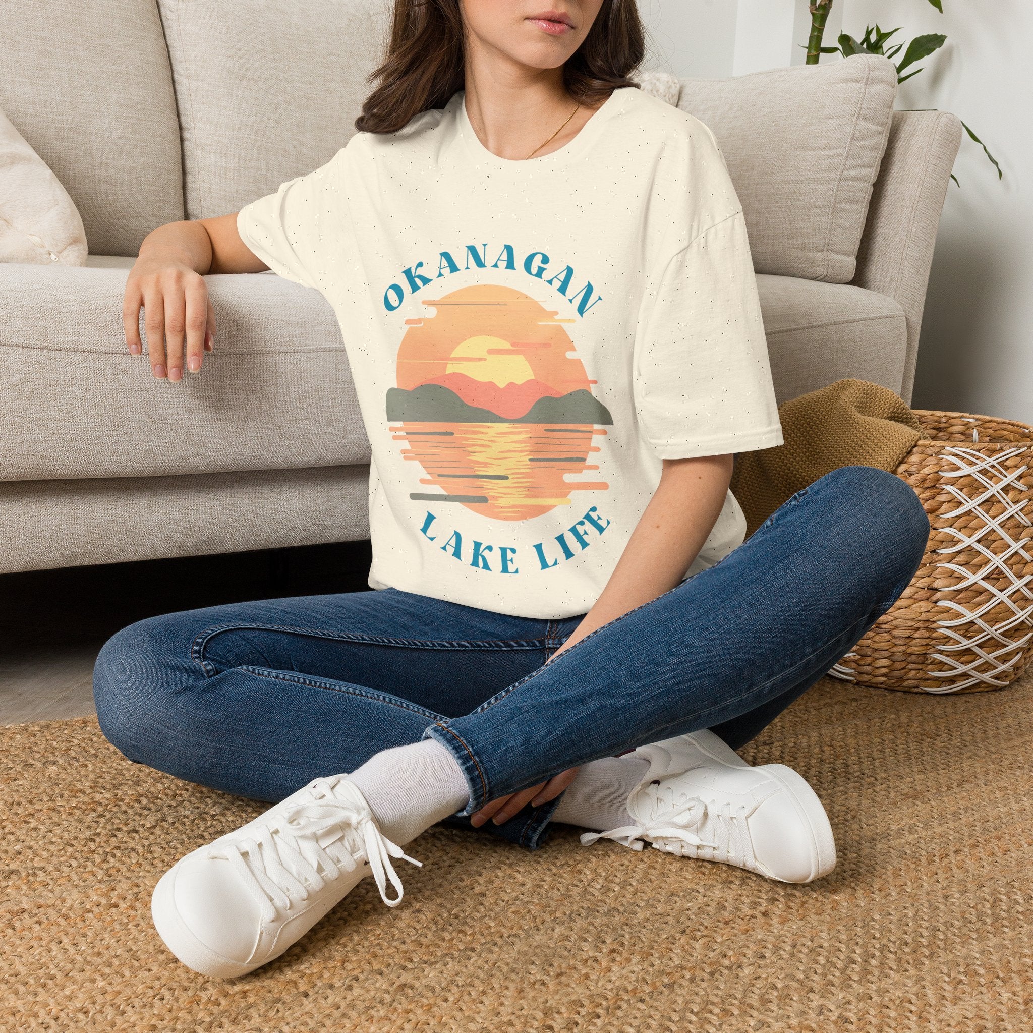 A woman sitting cross-legged on a woven rug in front of a light gray sofa, wearing a cream graphic T-shirt with a sunset lake design and the words 'OKANAGAN LAKE LIFE' in blue, paired with blue jeans and white sneakers.