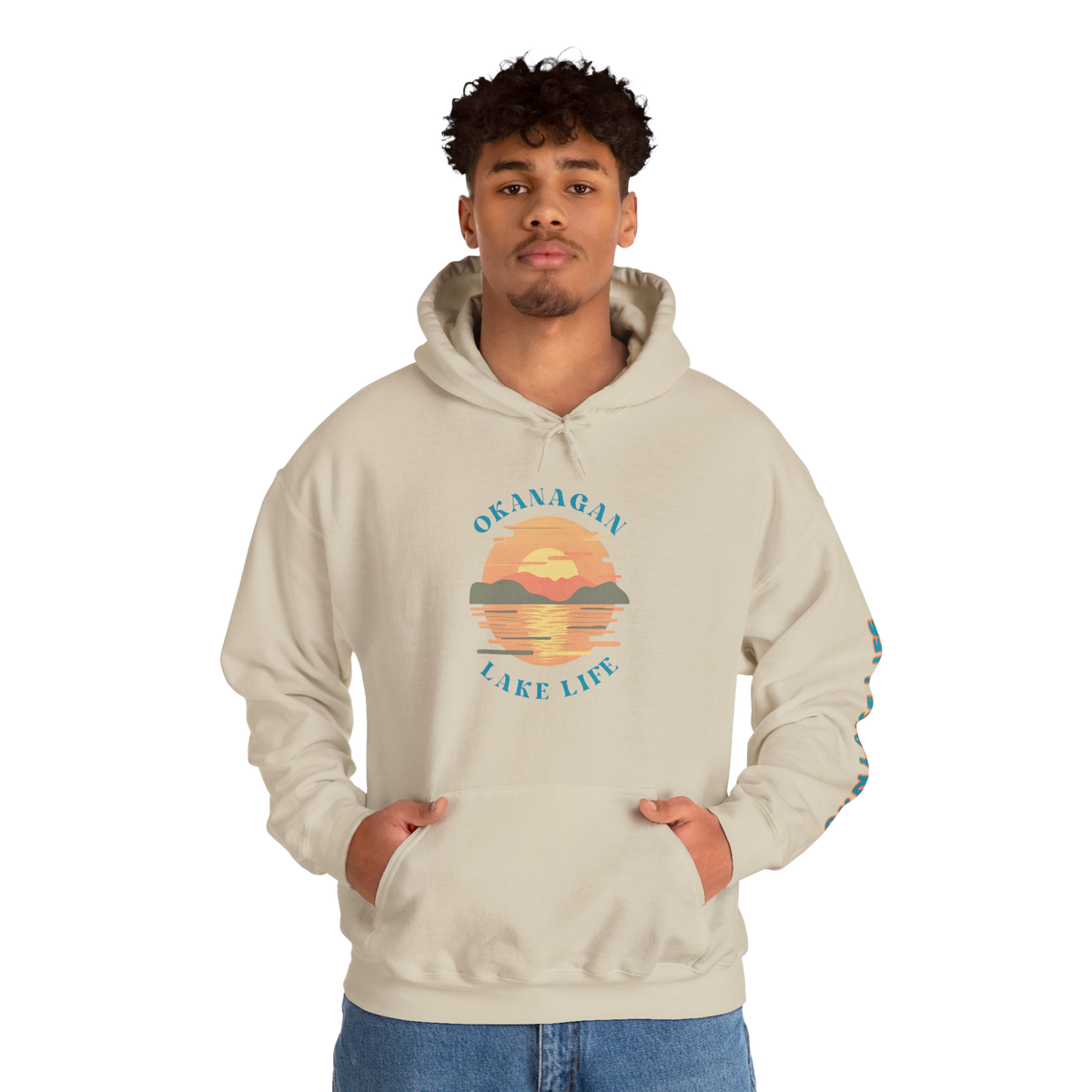 A man wearing a light beige pullover hoodie with a colorful 'Okanagan Lake Life' sunset graphic on the chest, posed against a solid white background.